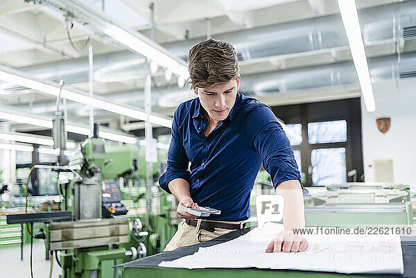 Male engineer examining blueprint while holding machine part in in industry