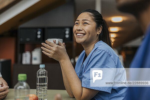 Smiling female healthcare worker holding cup while looking away sitting in hospital cafeteria
