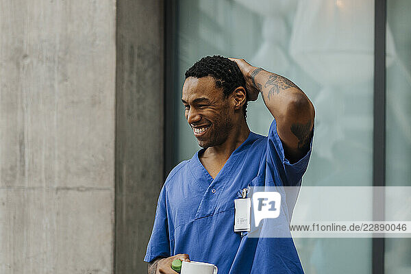 Happy male nurse holding mug with hand behind head standing in clinic