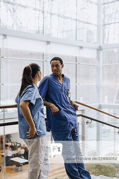 Male healthcare professional leaning on railing while talking with female coworker in hospital corridor