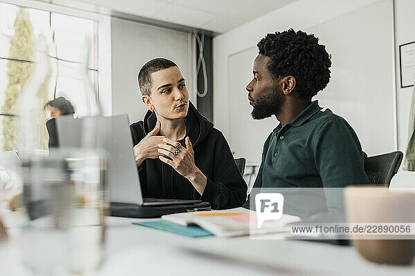 Non-binary entrepreneur discussing with male colleague while sitting at desk in tech office