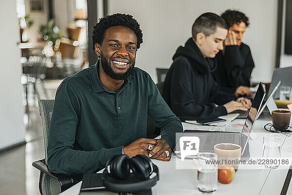 Portrait of smiling young businessman sitting with laptop in tech startup office