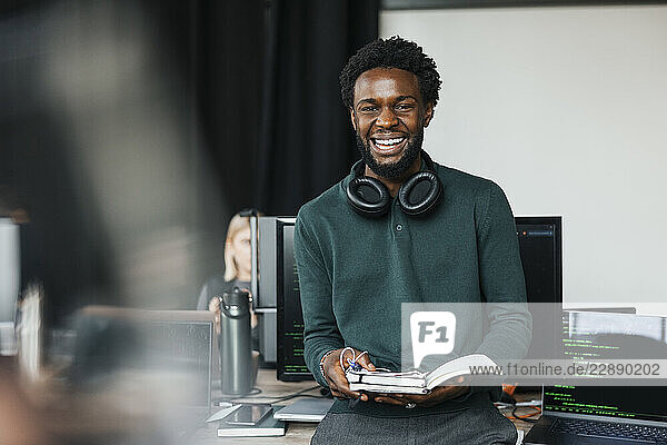 Portrait of happy young male IT professional with wireless headphones around neck holding diary while leaning at desk in