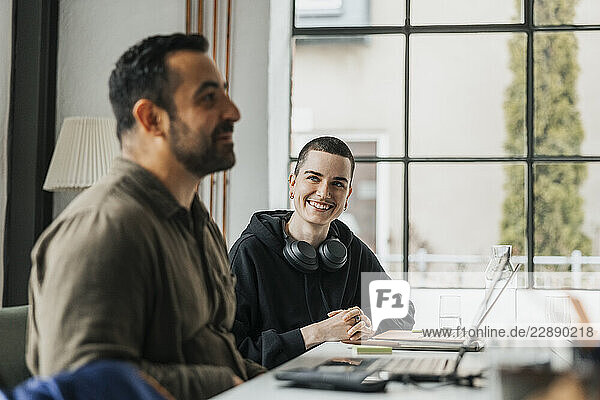 Happy non-binary software engineer sitting with male colleague at desk in tech start-up company