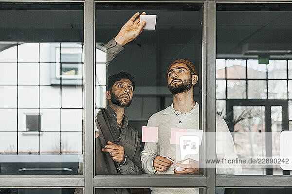 Male entrepreneurs sticking adhesive notes on glass window