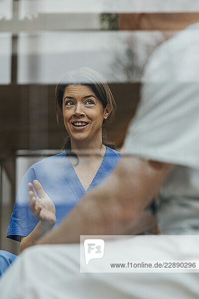 Smiling female medical professional gesturing while discussing with coworker seen through glass in clinic