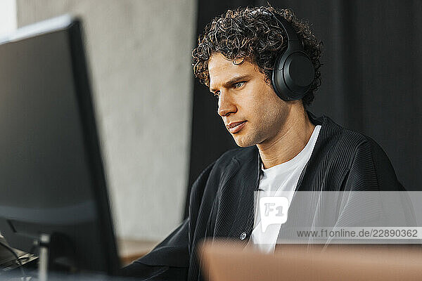 Curly haired young male computer programmer wearing wireless headphones using desktop PC in startup office