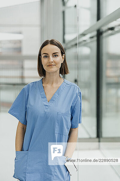 Portrait of short haired female medical expert leaning on glass wall with hands in pockets