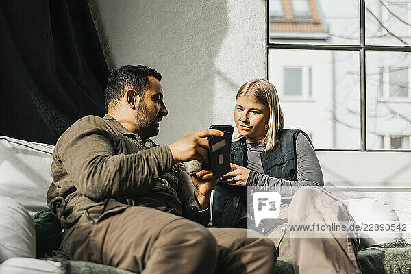Businessman showing smart phone while talking with female coworker sitting on sofa at tech startup office