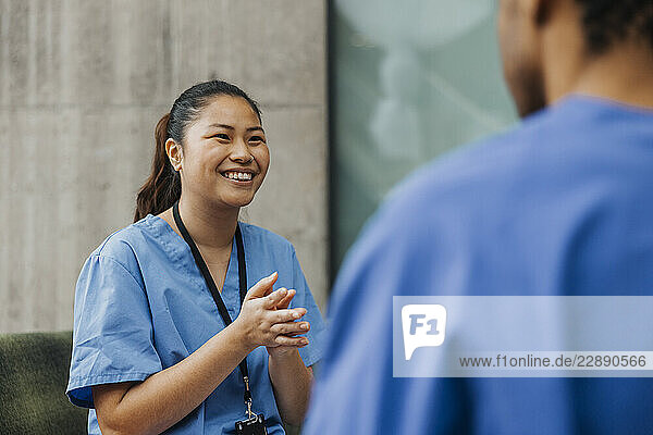 Smiling young female medical professional with hands clasped talking with colleague in clinic