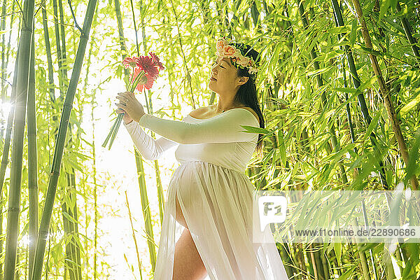 Pregnant woman in a white dress holding flowers in a sunlit bamboo forest. WA  USA