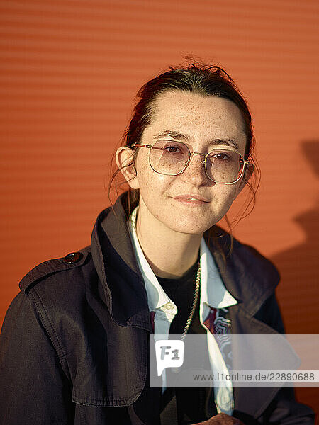 Young woman with glasses in front of an orange textured wall smiling in sunlight.
