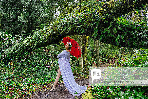Pregnant woman in white dress with red umbrella under a lush forest archway. WA  USA