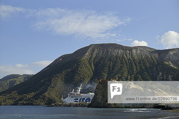 A ferry navigates near a volcanic island with lush green slopes and clear blue skies. Sicily  Italy