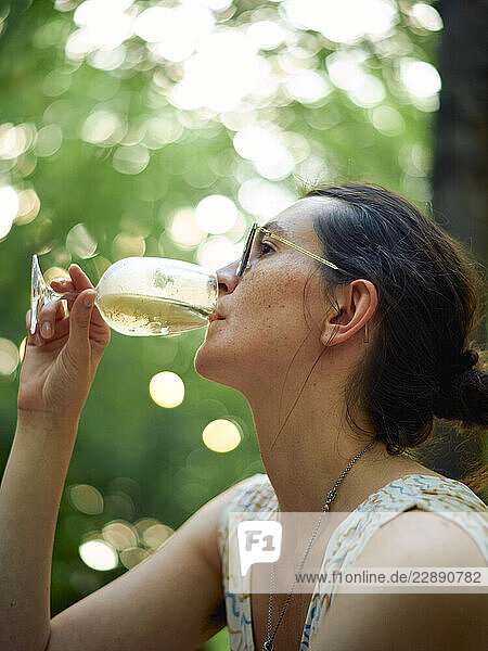 Woman sipping a drink from a wine glass outdoors with a blurred green background.