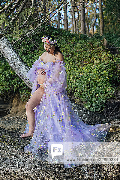 Pregnant woman in floral gown sits on a tree with lush greenery in the background. WA  USA