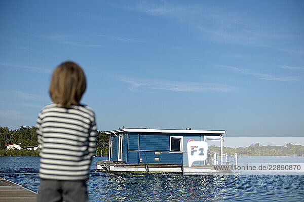Child in striped shirt watches a blue houseboat on a calm lake under a clear sky. Mecklenburg Lake Plateau  Mecklenburg-Western Pomerania  Germany
