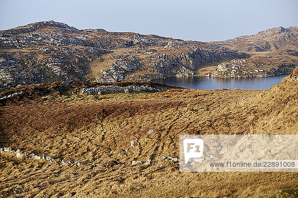 Rugged brown hills and rocky terrain surrounding a serene blue lake under a clear sky. West coast Ireland