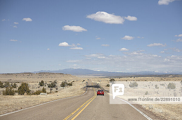 A red car on an empty road stretches toward distant mountains under a clear blue sky. Road Trip USA
