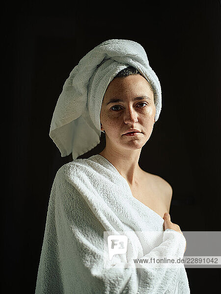 Woman wrapped in towel with a towel on head against a dark background.