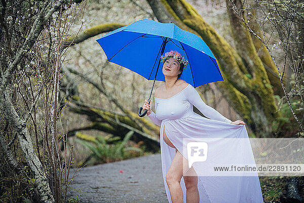 Pregnant woman in flowy white dress holds blue umbrella on forest path with pink flower petals. WA  USA