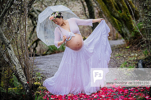 Pregnant woman in flowy white dress holds umbrella on forest path with pink flower petals. WA  USA