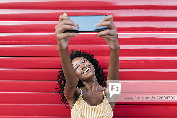 Young woman laughing while taking selfie through mobile phone in front of red shutter
