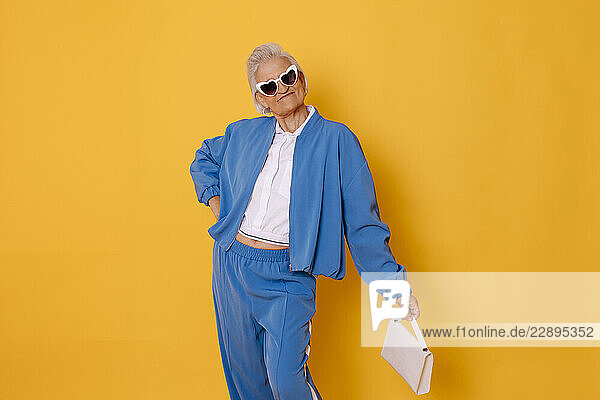 Stylish woman in blue outfit posing with sunglasses and handbag against a yellow background