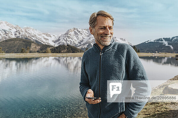 Smiling mature man holding mobile phone while standing against Prinzensee  Salzburger Land  Austria