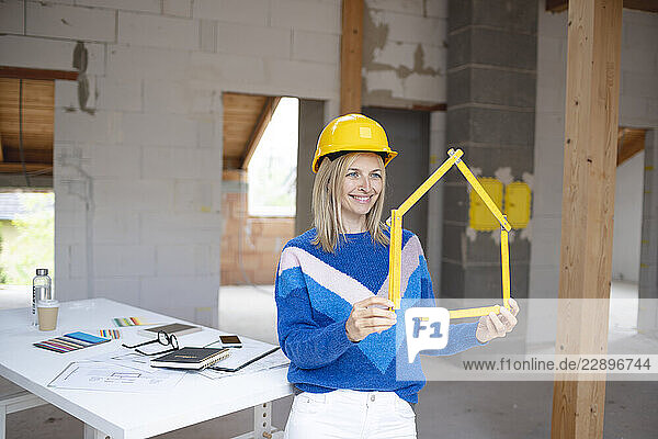 Smiling female building contractor in hardhat making house shape with folding ruler at apartment