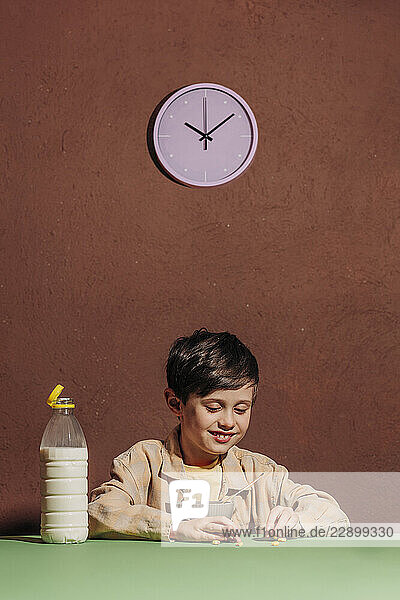Boy eating breakfast cereal with milk at a table under a purple clock