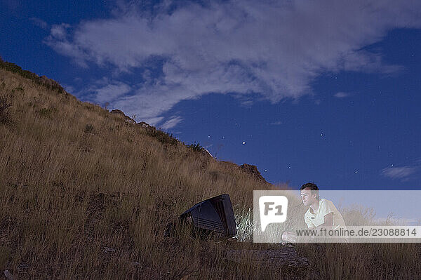 A person watches a glowing television in the middle of a vast barren field at dusk.